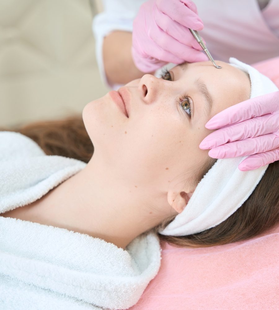 Cosmetologist cleans girl forehead with uno spoon in beauty saloon